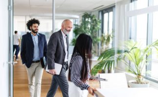 Expert Facilitation A group of business professionals, including a man with a beard and another with curly hair, walk through a bright corporate office surrounded by large windows and plants. The scene conveys teamwork and professionalism.